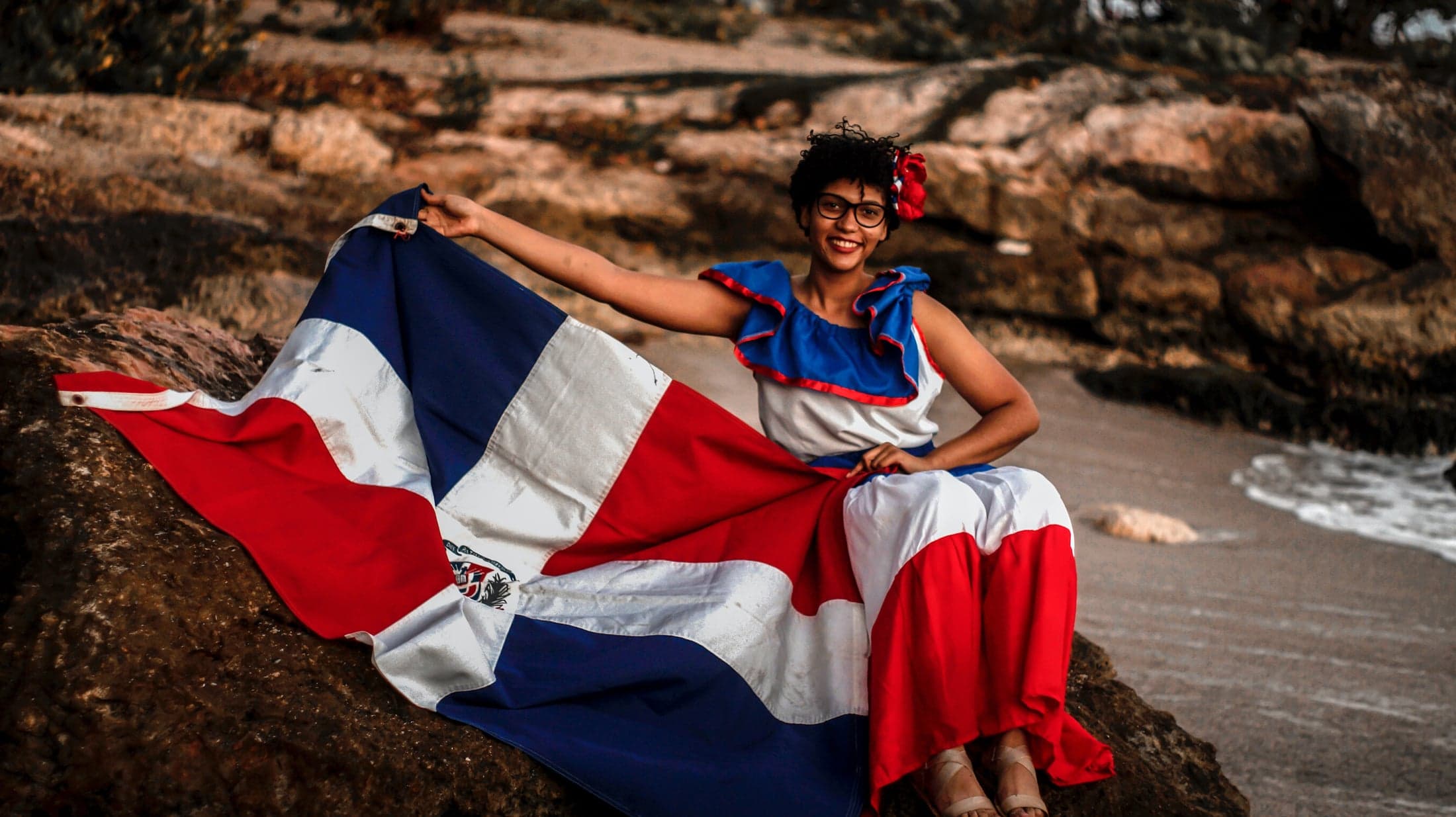 Mujer con traje típico sosteniendo la bandera dominicana junto al mar